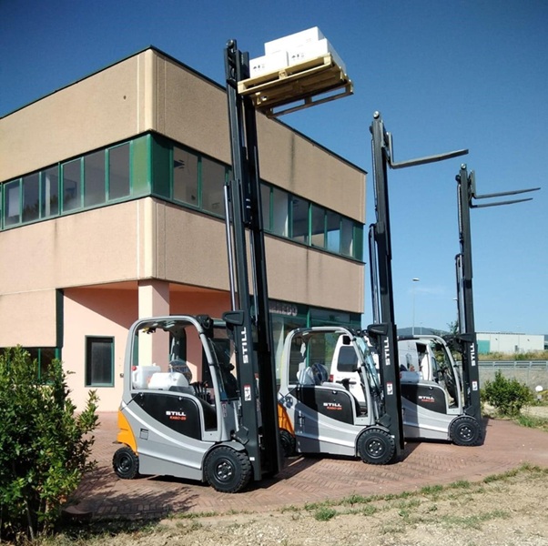 Tre carrelli elevatori STILL grigi e neri, uno con pallet di scatole bianche sollevato, davanti a un edificio beige